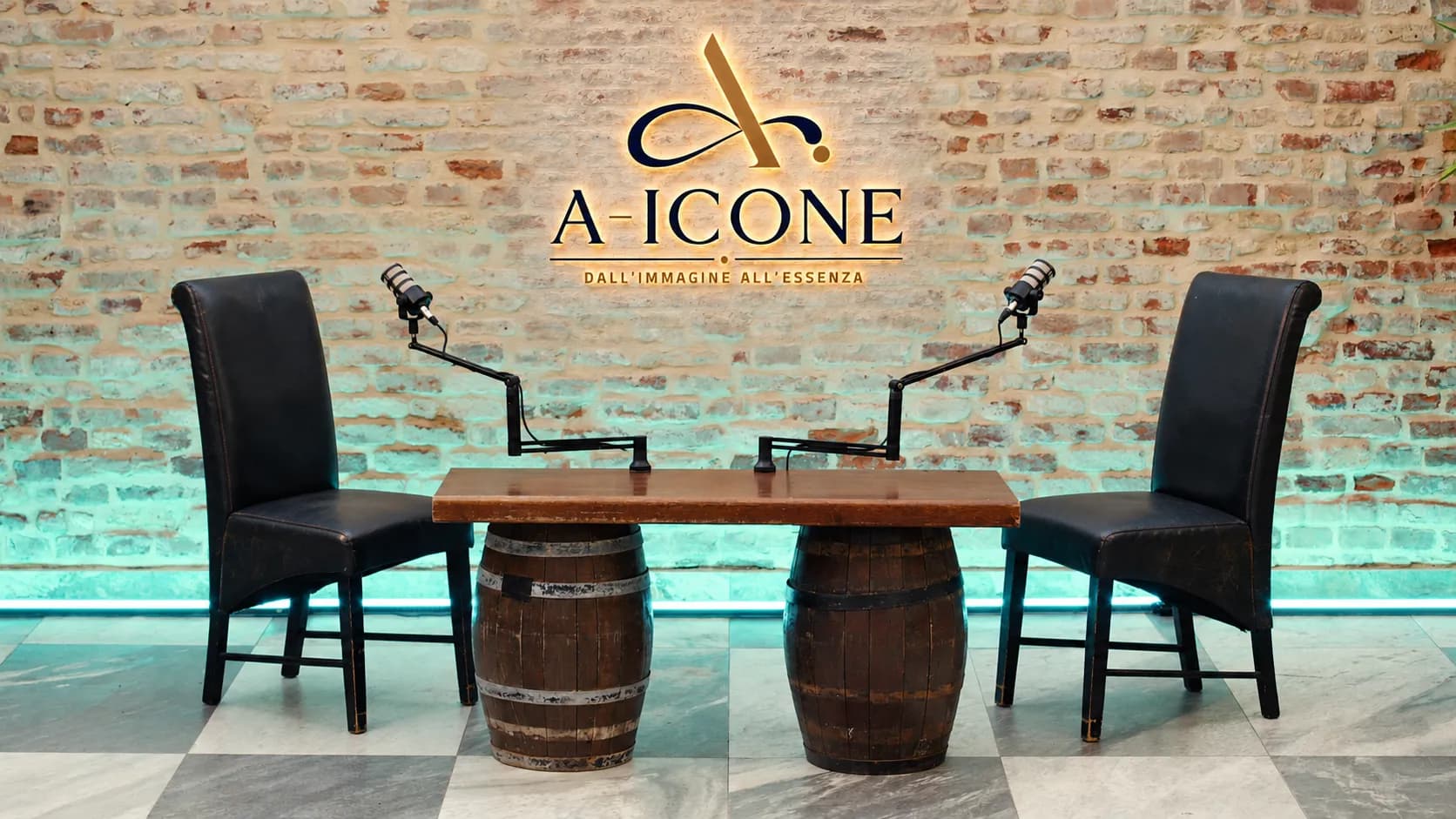 Il set di A-Icone: due poltrone e due microfoni ai lati di un tavolo su botti, sotto l’insegna luminosa.